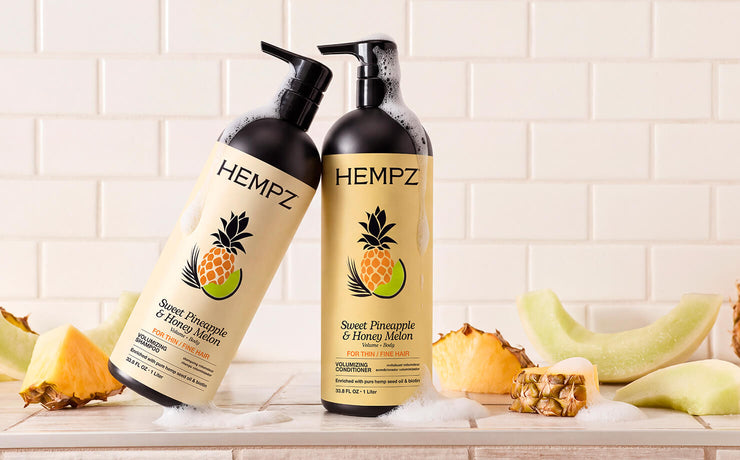 Two pump bottles of HEMPZ Sweet Pineapple & Honey Woolen hair care products standing on a light stone surface with pineapple and melon slices around them, against a white subway tile wall.