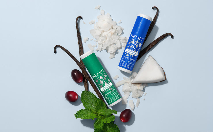Two tubes of Hempz lip balm, one green and one blue, styled with vanilla beans, coconut, dried cranberries, and mint leaves on a light blue background.