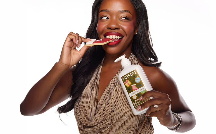 A smiling woman with long dark hair eating a sugar cookie while holding a white pump bottle labeled HEMPZ Vanilla Sugar Cookie bottle. She is wearing a textured beige top against a plain white background.